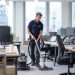 a male professional carpet cleaner in a small modern London IT startup office, steam-cleaning a grey carpet